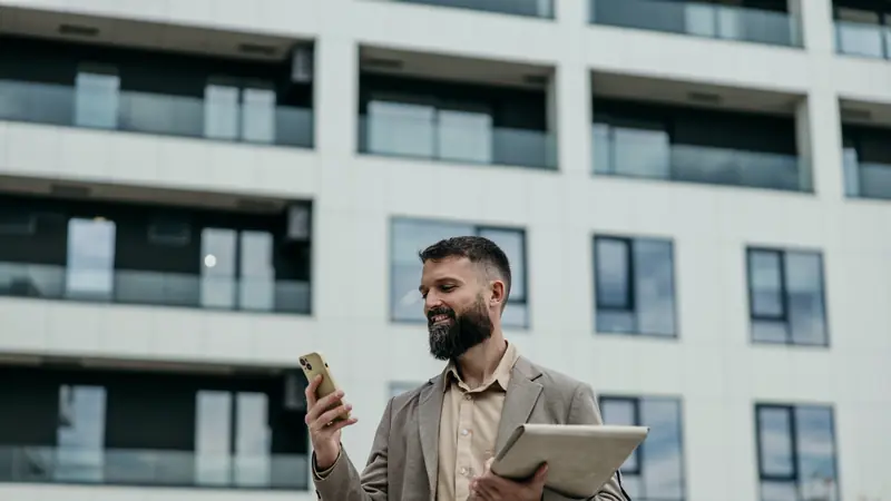 Immobilienmakler mit Smartphone und Mappe vor einem Wohngebäude